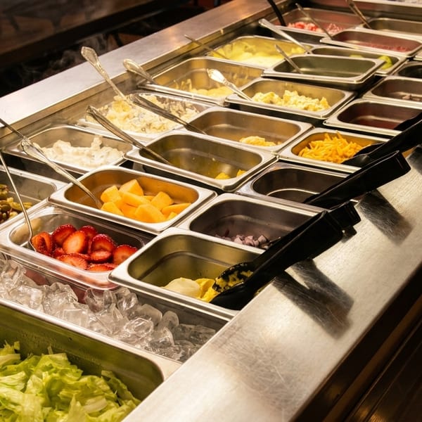 A salad bar with various toppings like lettuce, strawberries, fruit, and cheese in metal containers.