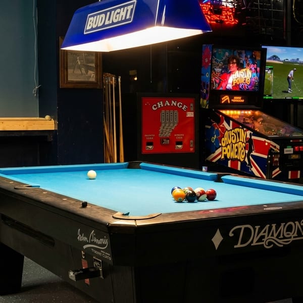 A pool table with blue felt and billiard balls, surrounded by arcade games and a Bud Light sign.