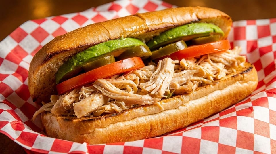Shredded chicken sandwich with avocado, tomatoes, and jalapeños on a toasted bun in a red checkered basket.