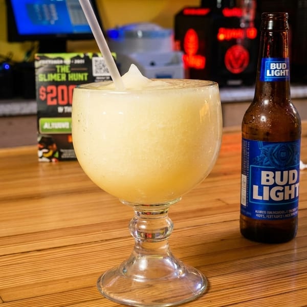 A frozen margarita with a straw and a bottle of Bud Light on a wooden bar.
