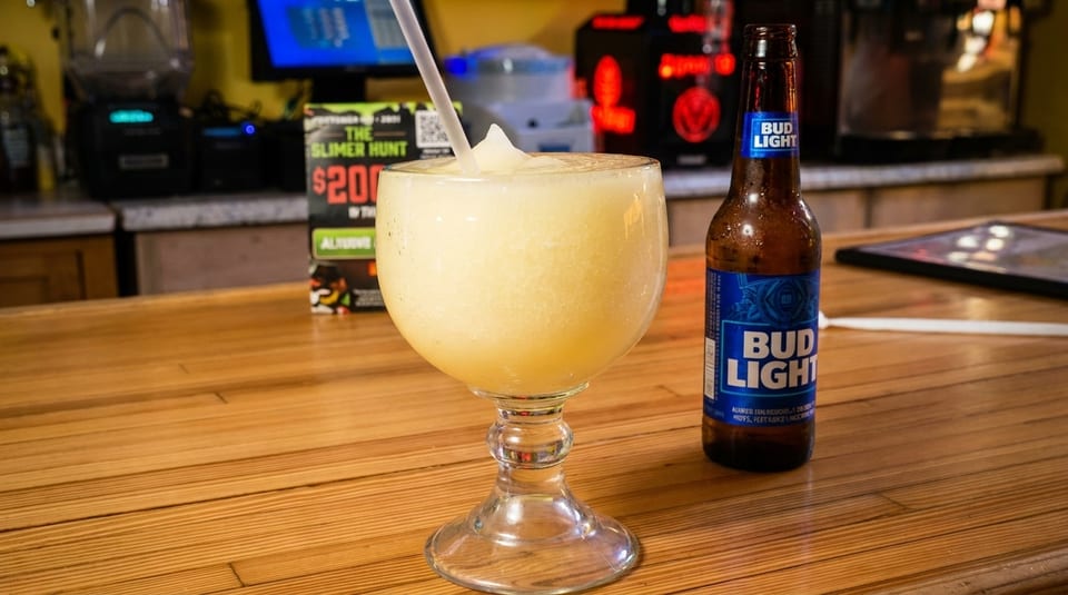 A frozen margarita with a straw and a bottle of Bud Light on a wooden bar.