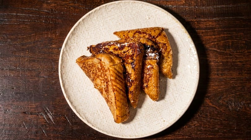 Golden brown French toast slices dusted with powdered sugar on a white plate, on a dark wooden table.