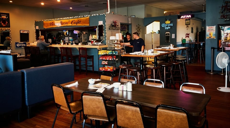 A diner interior with tables, booths, a bar, and two employees behind a snack display.
