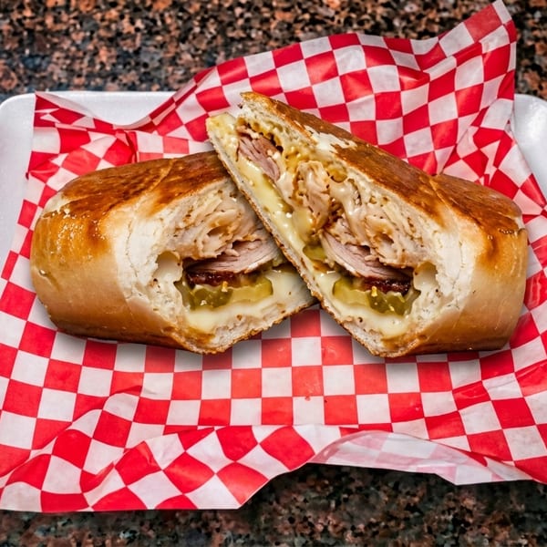 A toasted Cuban sandwich, cut in half, revealing turkey, ham, pickles, and melted cheese on checkered paper.