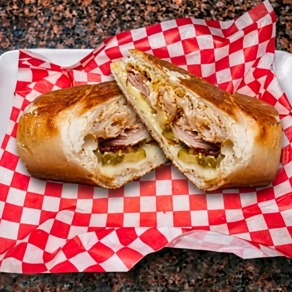 A toasted Cuban sandwich, cut in half, revealing turkey, ham, pickles, and melted cheese on checkered paper.
