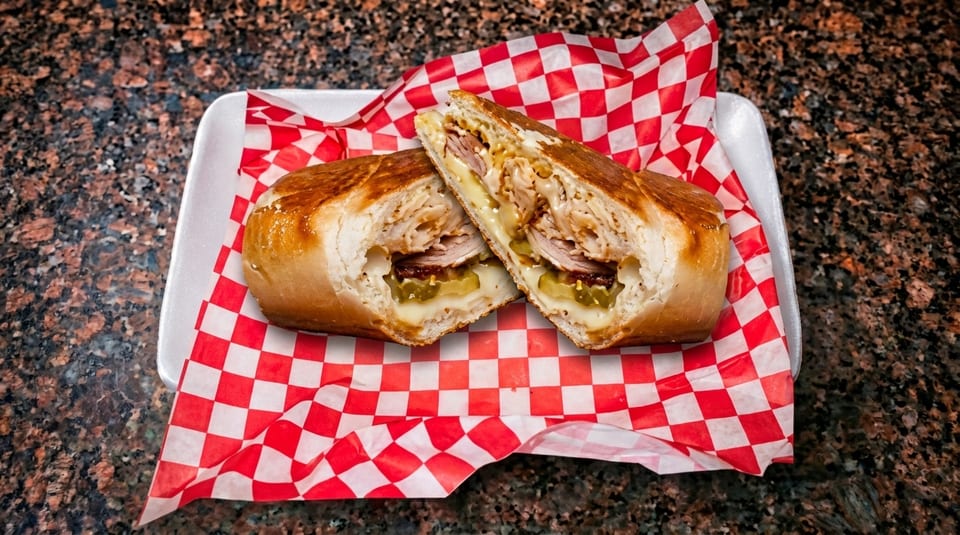 A toasted Cuban sandwich, cut in half, revealing turkey, ham, pickles, and melted cheese on checkered paper.